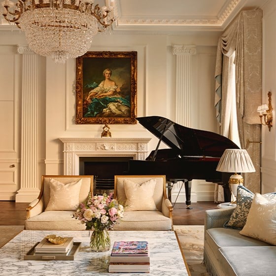 Elegant lounge with crystal chandelier, grand piano, classical painting above the fireplace, and a marble coffee table adorned with fresh flowers.