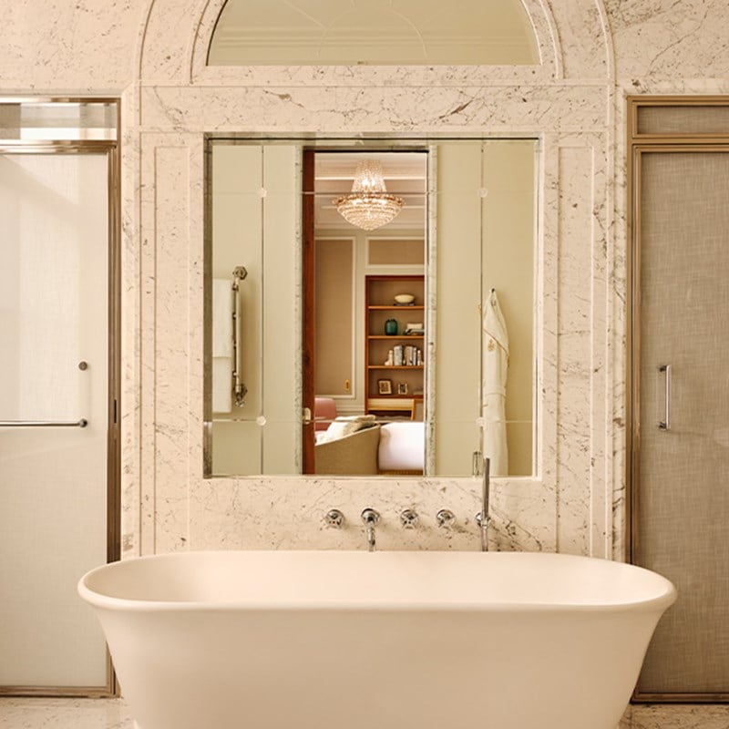 Freestanding white bathtub set against a marble wall with mirror reflecting a chandelier and softly lit interior of the suite.