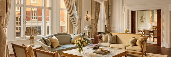 Refined drawing room with soft gold and blue sofas, large windows, floral arrangements, and glimpse into the adjoining formal dining room.
