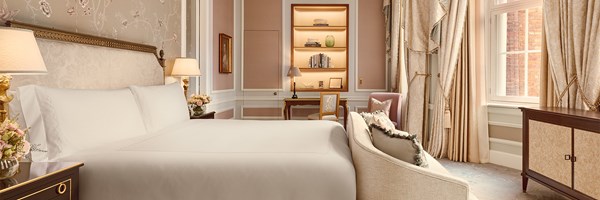 Bedroom with grand upholstered headboard, crisp white bedding, floral wallpaper, writing desk, and richly draped windows with views of red-brick façades.