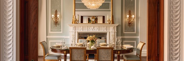 Ornate dining room with intricately carved white fireplace, pastel green walls, gold sconces, and glass-topped table beautifully set for six.
