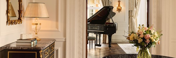 A luxurious sitting room with a marble table, fresh flowers, and a black Steinway grand piano framed by tall curtains.