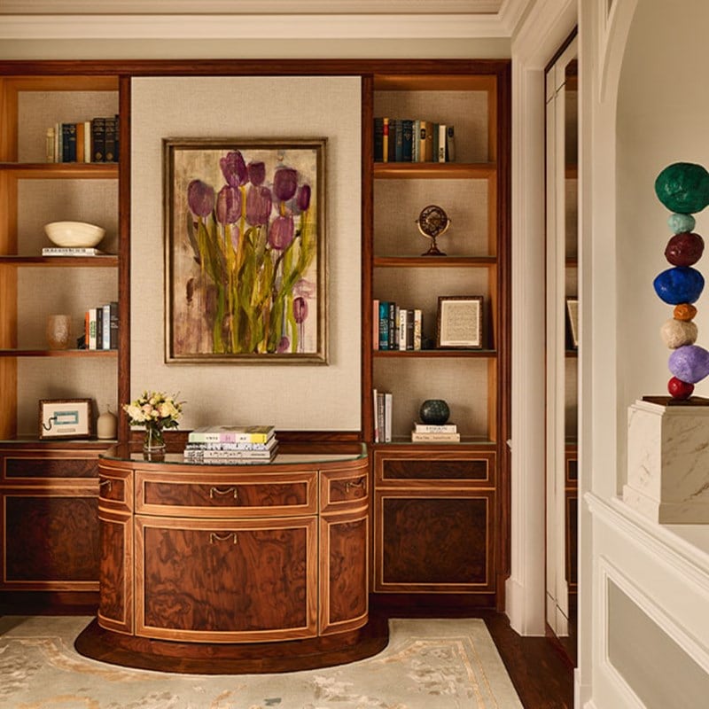 A library alcove with built-in bookshelves, floral artwork, and a polished wood writing desk.
