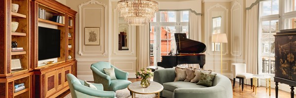Sophisticated living room with pastel armchairs, a curved sofa, grand piano, and crystal chandelier under high ceilings and tall windows.