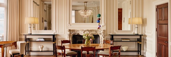 Refined dining space with a round wooden table, ornate marble fireplace, large mirrors, and soft cream wall panelling.