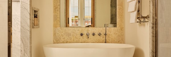 Grand Terrace Suite bathroom with a freestanding oval bathtub, marble walls, and a large mirror reflecting French doors and natural light.