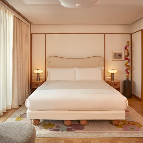 A front-facing view of a king-size bed with crisp white linens, positioned on a floral rug and flanked by bedside tables and Art Deco lamps.