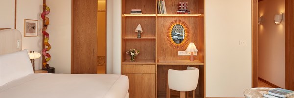 View of a wooden built-in desk and shelving with a sculptural mirror and books, next to the bed and colourful sculptural floor lamp.