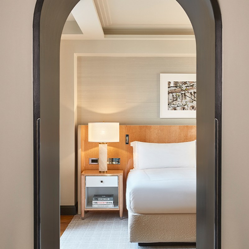 View through an arched doorway into a softly lit bedroom with a wooden headboard, white bed linens, and a bedside table with lamp.