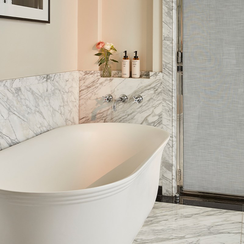 Freestanding bathtub with chrome fixtures and marble surroundings, complemented by fresh flowers and toiletries on a recessed shelf.