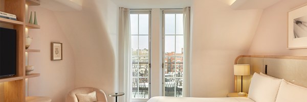 Bedroom view looking out through tall French windows to a private balcony, with cream curtains and pastel-toned interior.