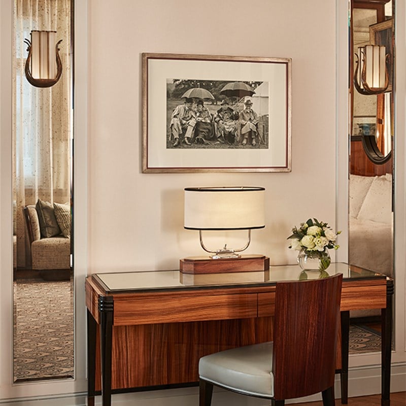 Glass-topped wooden writing desk with a contemporary lamp, chair, and vase of white flowers, framed by mirrors and artwork on the wall.