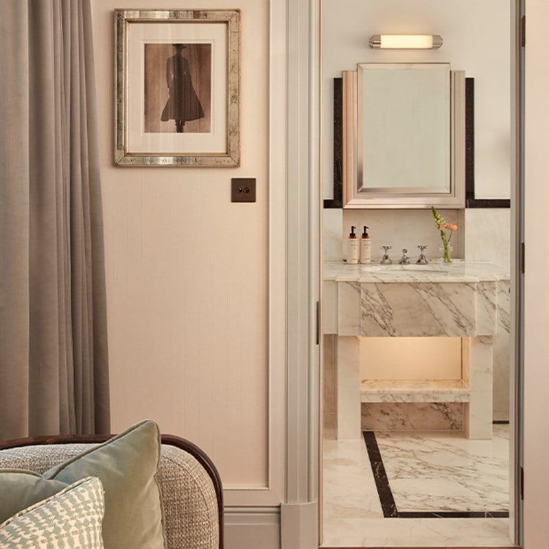View into a marble bathroom with a vanity, mirror, and framed fashion artwork on the outer wall.