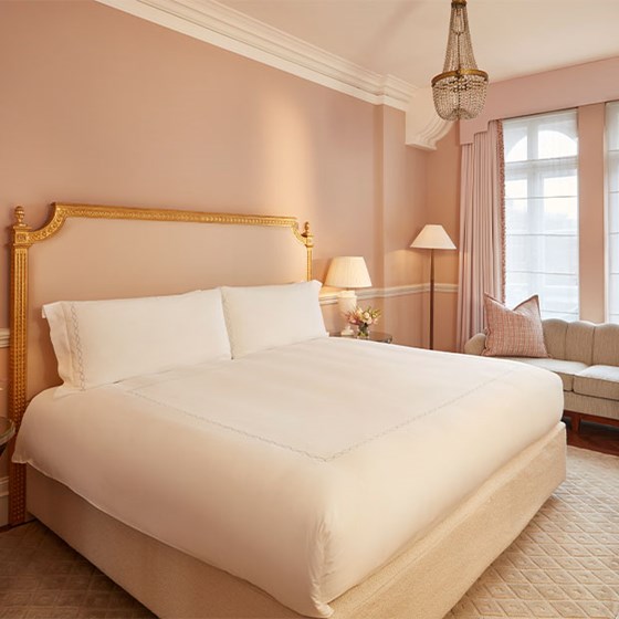 Softly lit Mayfair Room with blush pink walls, a king-size bed with ornate gold headboard, classic chandelier, and a curved cream sofa beside tall windows dressed in sheer and pink curtains.