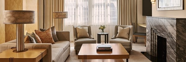 A spacious living area with a neutral-toned sofa, two armchairs, and a wooden coffee table beneath large sheer-draped windows, accented by soft lighting and a marble fireplace on the right.