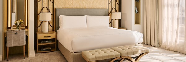 A serene bedroom featuring a large bed with crisp white linens, a padded bench at the foot, bedside tables with lamps, and full-length curtains framing floor-to-ceiling windows.
