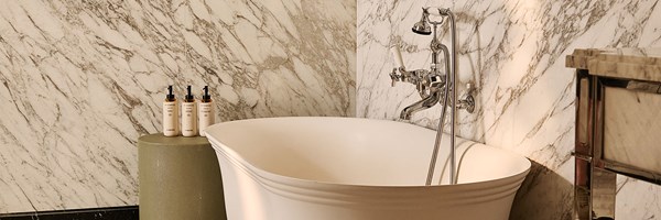 A marble-clad bathroom with a freestanding white bathtub, chrome fixtures, and luxury toiletries arranged on a small side table.