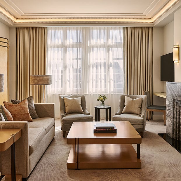 A bright living space with a neutral sofa, two armchairs, a wooden coffee table with books, and tall windows dressed with sheer curtains and drapes.