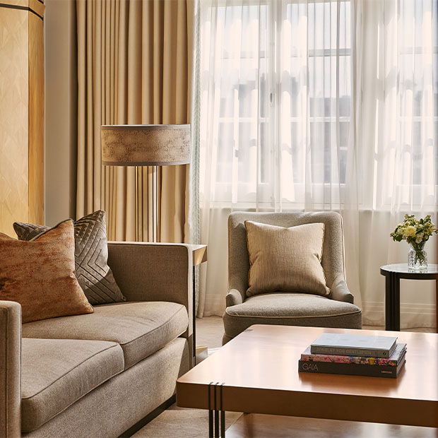 A close-up of a beige sofa with textured cushions beside an armchair positioned near tall curtained windows and a small floral arrangement.