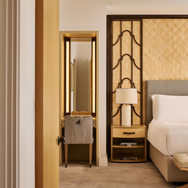 A partial view of the bedroom showing a dressing table with an upholstered chair and illuminated mirrors beside the bed with a sculpted wooden headboard.