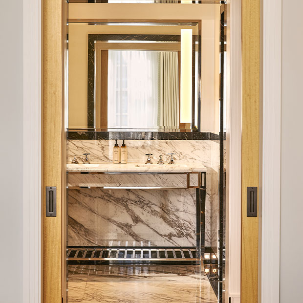 A marble bathroom vanity with twin sinks reflected through an open doorway, styled with luxury toiletries and chrome fixtures.