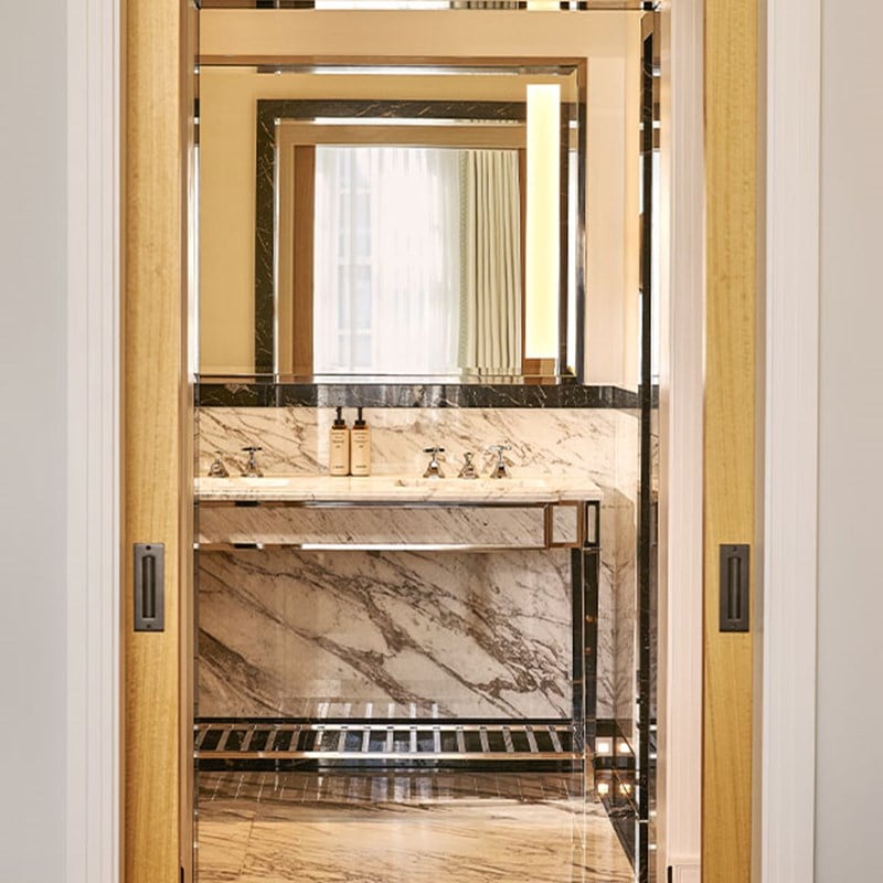 A marble bathroom vanity with twin sinks reflected through an open doorway, styled with luxury toiletries and chrome fixtures.