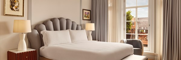 Bright bedroom with a king bed featuring a sculpted grey headboard, flanked by two white lamps on bedside tables, and a window with floor-length curtains opening to a terrace view.