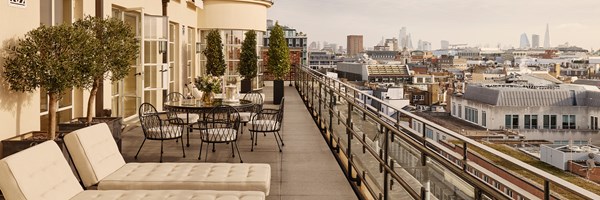 Expansive private terrace with sun loungers, black wrought iron dining set, and panoramic London skyline views in golden afternoon light.