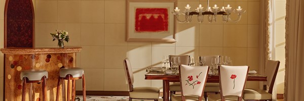 Dining area with a polished wooden table, cream embroidered chairs, chandelier, and a curved bar featuring a marquetry dot pattern.