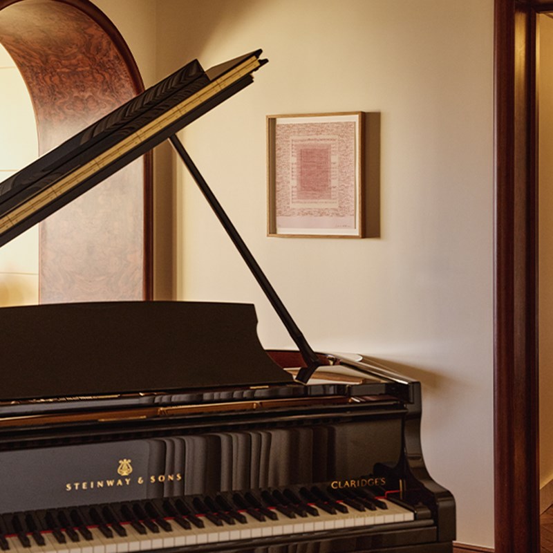Close-up of a black Steinway & Sons grand piano with “Claridge’s” inscription, positioned beneath a framed artwork and archway.
