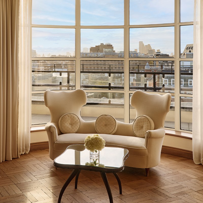 Curved cream high-back loveseat with round cushions and black coffee table, positioned beside bay windows overlooking the London cityscape.