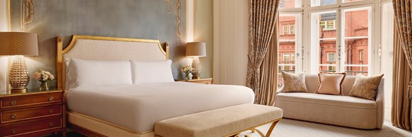 Elegant bedroom with a king-size bed, golden frame, fluted lamps, and floral drapes overlooking red-brick architecture.