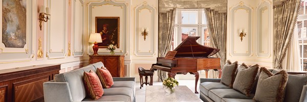 Opulent living room with two powder-blue sofas, carved wood grand piano, gilded wall panels, and tall curtained windows.