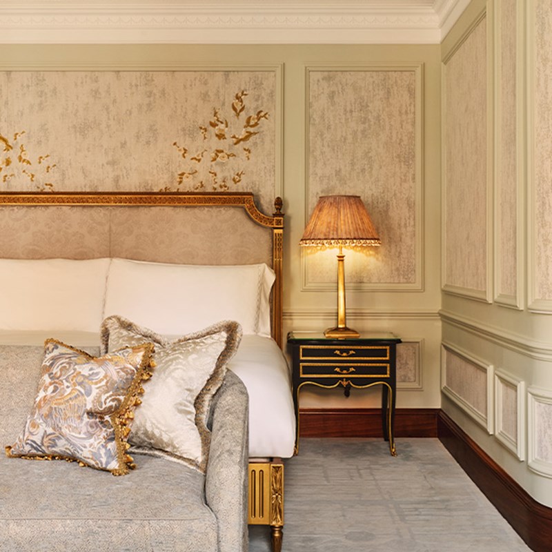Close-up of a Brook Suite bed at Claridge’s, featuring an ornate gold-trimmed headboard, embroidered wall panels, and a plush chaise with tasseled pillows, beside a black and gold bedside table with a pleated lampshade.