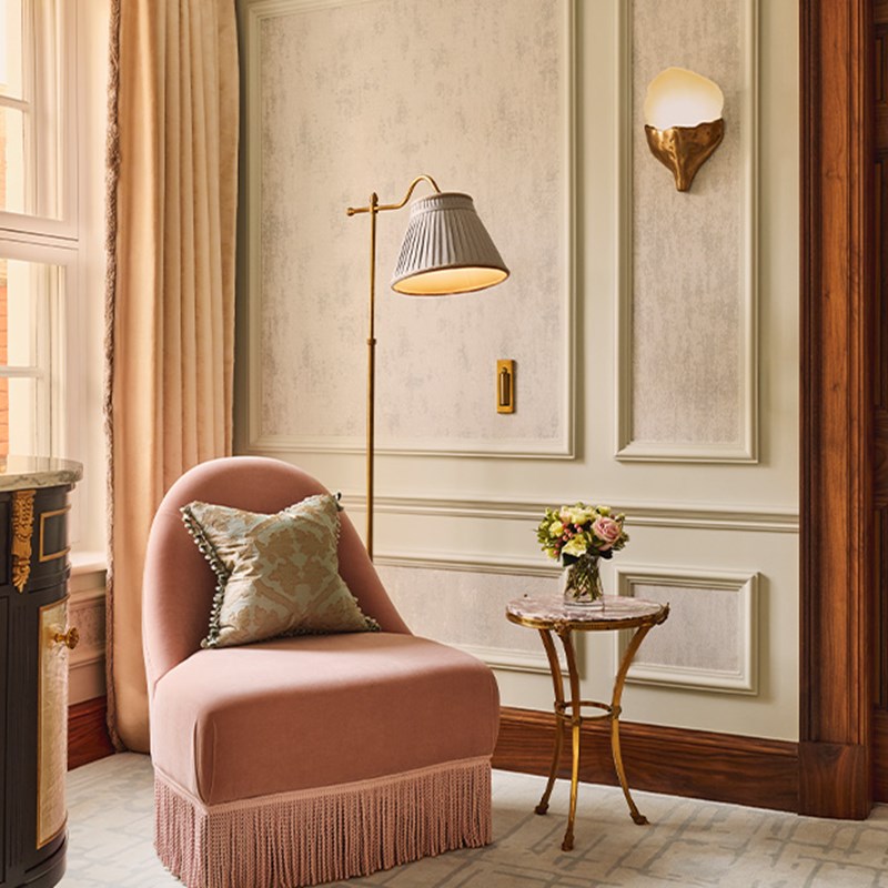 Reading corner in the Brook Suite at Claridge’s, with a fringed pink velvet chair, a gold floor lamp, a round marble-topped side table with a vase of flowers, and pale decorative wall panels.