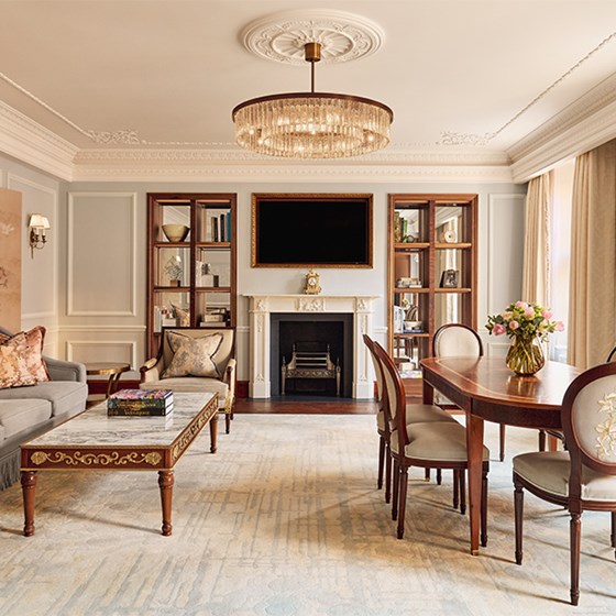 Elegant living and dining space in the Brook Suite at Claridge’s, with a grey sofa, carved marble fireplace, crystal chandelier, and large wooden bookshelves, combining traditional and modern elements.