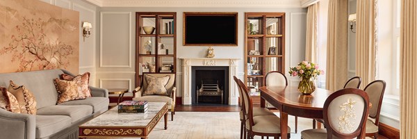 Panoramic view of the Brook Suite’s lounge and dining area at Claridge’s, with a pale blue and cream palette, crystal chandelier, ornate fireplace, and wooden dining table surrounded by floral-embroidered chairs.