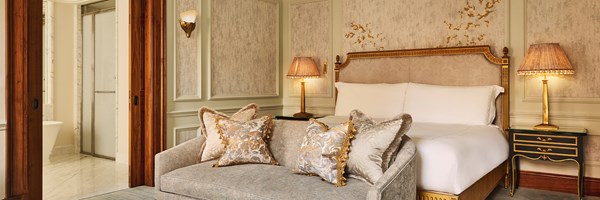 Wide-angle view of the Brook Suite bedroom at Claridge’s, featuring a gilded headboard, plush silver sofa, twin black lacquered bedside tables with gold lamps, and a view into the marble bathroom.