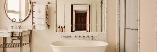 Brook Suite bathroom at Claridge’s, with a freestanding white bathtub, twin marble-topped vanities, walk-in shower, and mirrored wall reflecting the suite’s wooden doors and corridor.