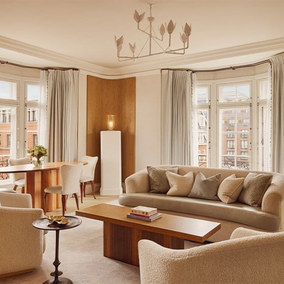 Bright and airy living area with curved cream sofa, wood coffee table, and dining set by tall windows with pale curtains in Claridge's Terrace Suite.