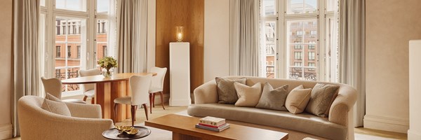 Spacious open-plan living and dining area with cream-toned seating, light wood furnishings, and multiple French windows offering views of red-brick buildings.