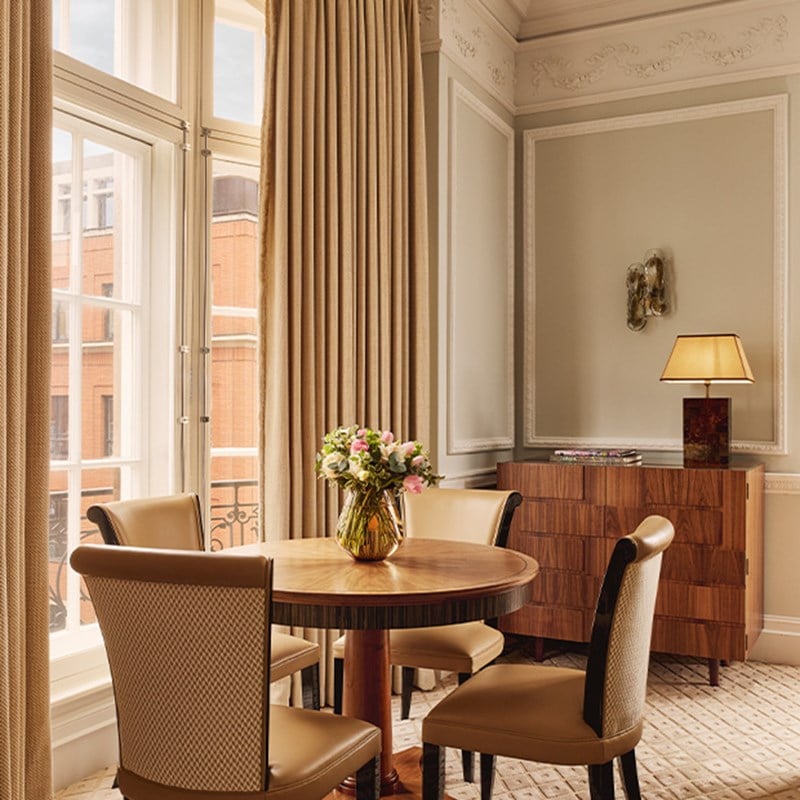Intimate dining area with round wood table and four cream leather chairs, set beside tall windows with beige curtains and a polished chest of drawers.