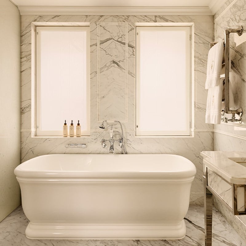Marble bathroom with freestanding white bathtub, twin frosted windows, and chrome fixtures, with Claridge’s toiletries neatly arranged on the sill.
