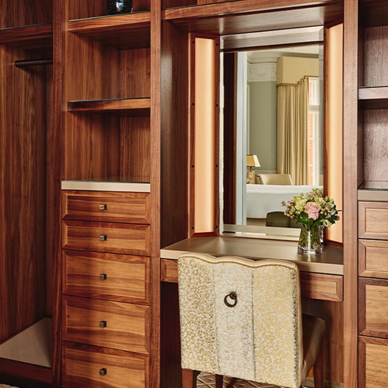 Walk-in wardrobe with open wooden shelving, built-in drawers, and a mirrored dressing table with an upholstered chair and fresh flowers.