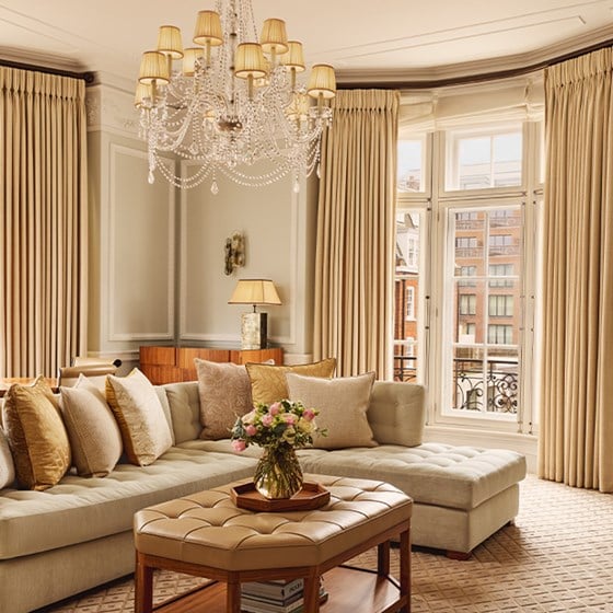 Elegant living room with L-shaped cream sofa, tufted ottoman, chandelier, and large windows framed by gold curtains offering views of Mayfair buildings.