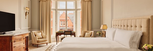 Tranquil bedroom with large upholstered bed, vintage-style desk by bay windows, and warm neutral furnishings accented by natural daylight.