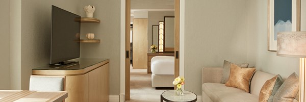 Contemporary living area with soft beige sofa, TV console, and view through sliding door into bedroom with large bed and lit vanity mirror.