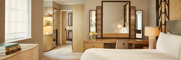 Spacious bedroom with Art Deco-inspired headboard, light wood desk with illuminated mirrors, and soft ambient lighting throughout.