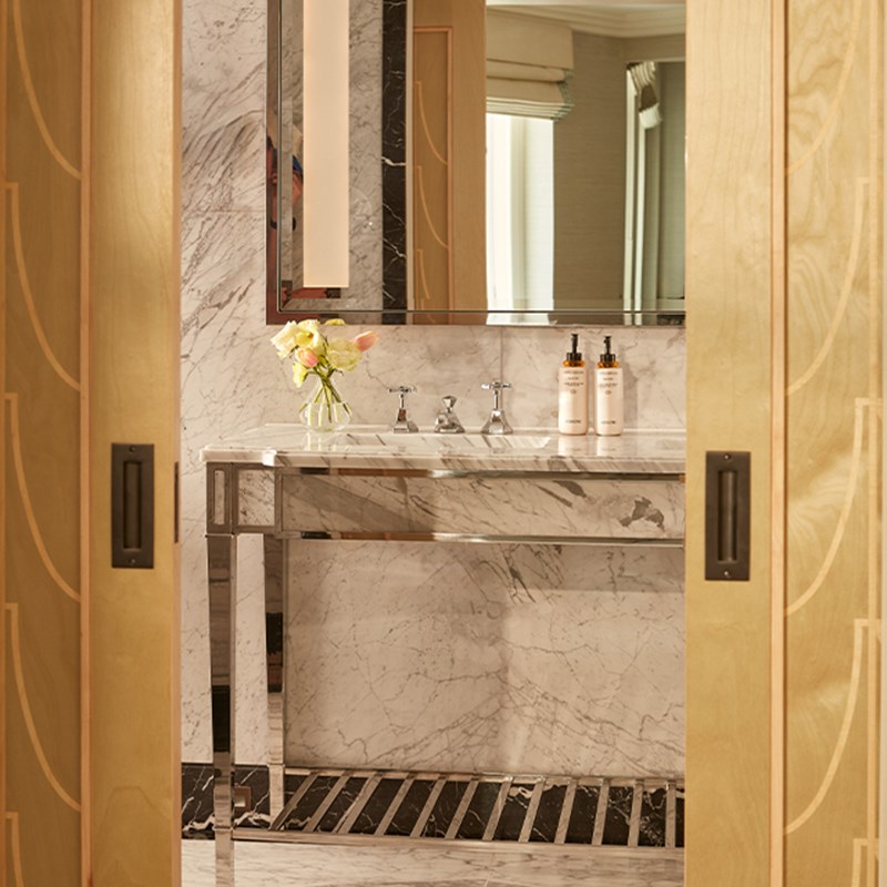 Marble bathroom with chrome vanity, large mirror, Claridge’s toiletries, and sliding wood doors with curved inlay detail.