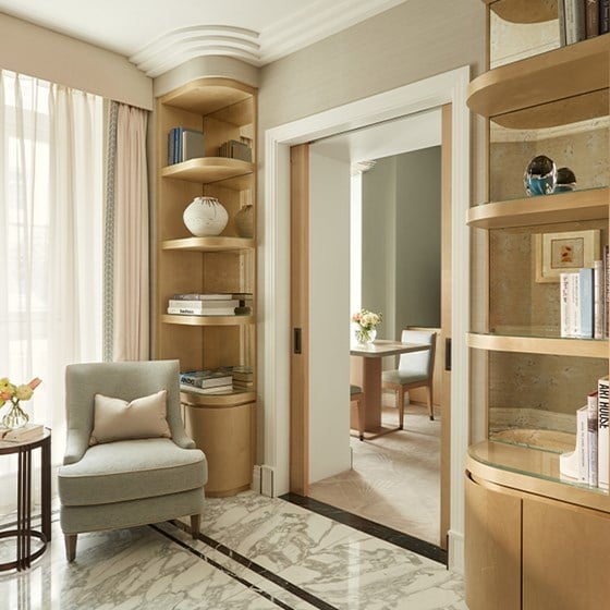 Sitting area with pale grey armchair, round side table, and built-in curved shelving filled with books and ceramics, leading to a dining nook.